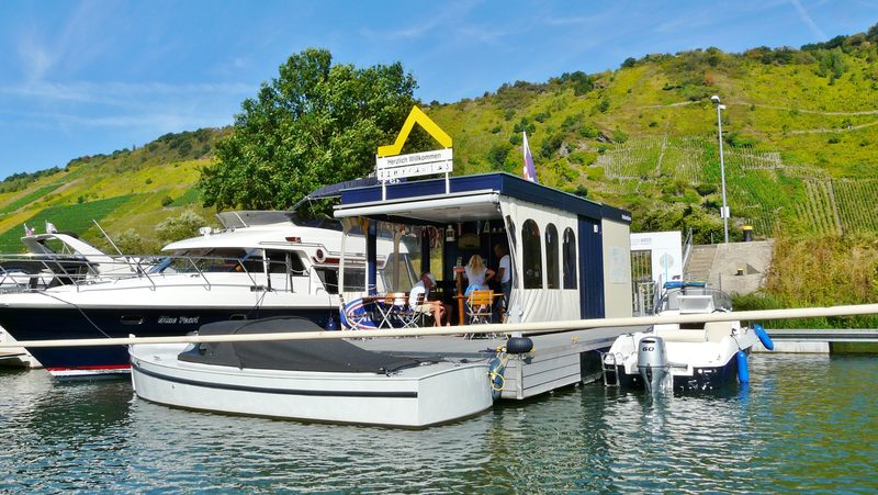 Schwimmende Hafenmeisterei des Yachthafens Traben-Trarbach mit Willkommensschild, davor Motorboote am Steg, dahinter der Weinberg.