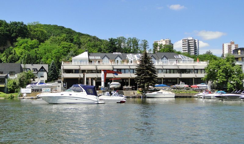 Die Steganlage des Wassersportzentrums Grühn von der Mosel aus gesehen, davor Motorboote am Schwimmsteg, dahinter Werfthalle und Kran, im Hintergrund ein bewaldeter Hang.