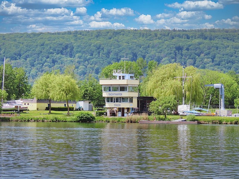 Der Regattaturm des Yachthafens Trier-Monaise vom Wasser aus gesehen, mit dem Schriftzug „Yachthafen Trier“, davor ein Schwimmsteg, rechts ein Kranausleger, dahinter ein bewaldeter Hang.